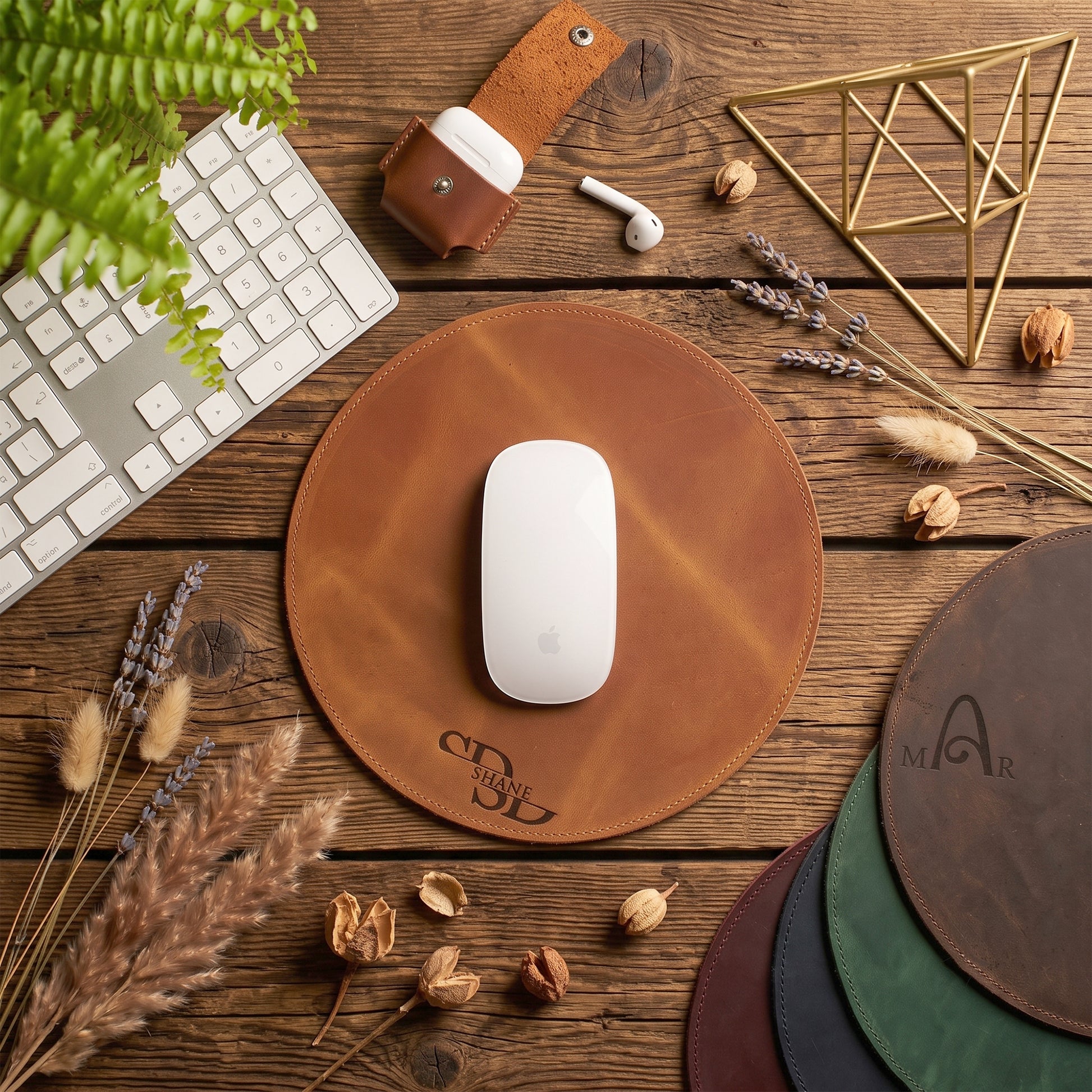 A round brown leather mouse pad on a rustic wooden desk, styled with a white computer mouse, keyboard, earbud case, dried flowers, and other colored mouse pads. The mouse pad has an embossed logo reading 'C & B Design Studio'.