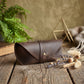 A dark brown leather eyeglasses case with a flap closure and a gold-tone snap button, resting on a rustic wooden table decorated with dried wheat, lavender, and a green fern in the background.