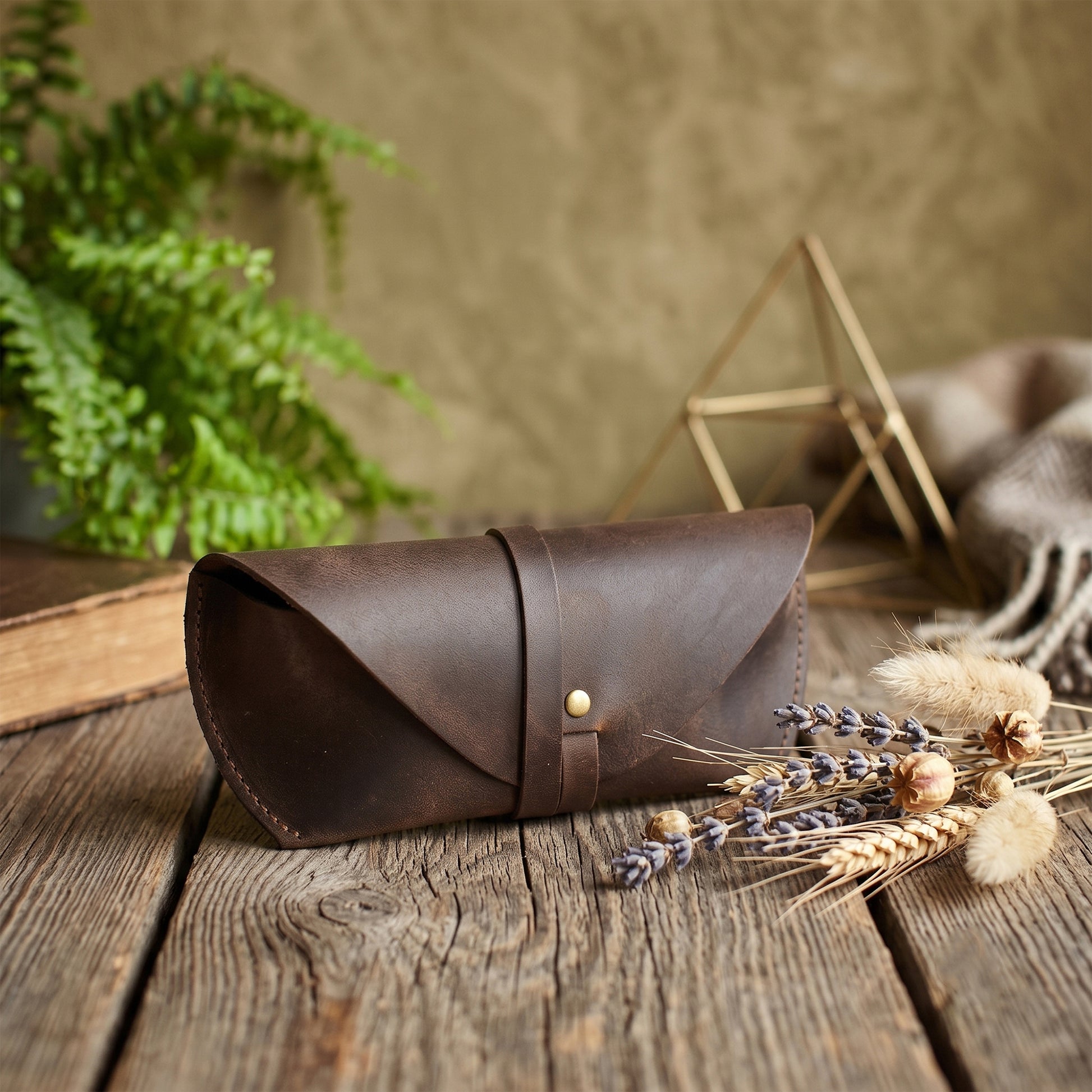 A dark brown leather eyeglasses case with a flap closure and a gold-tone snap button, resting on a rustic wooden table decorated with dried wheat, lavender, and a green fern in the background.