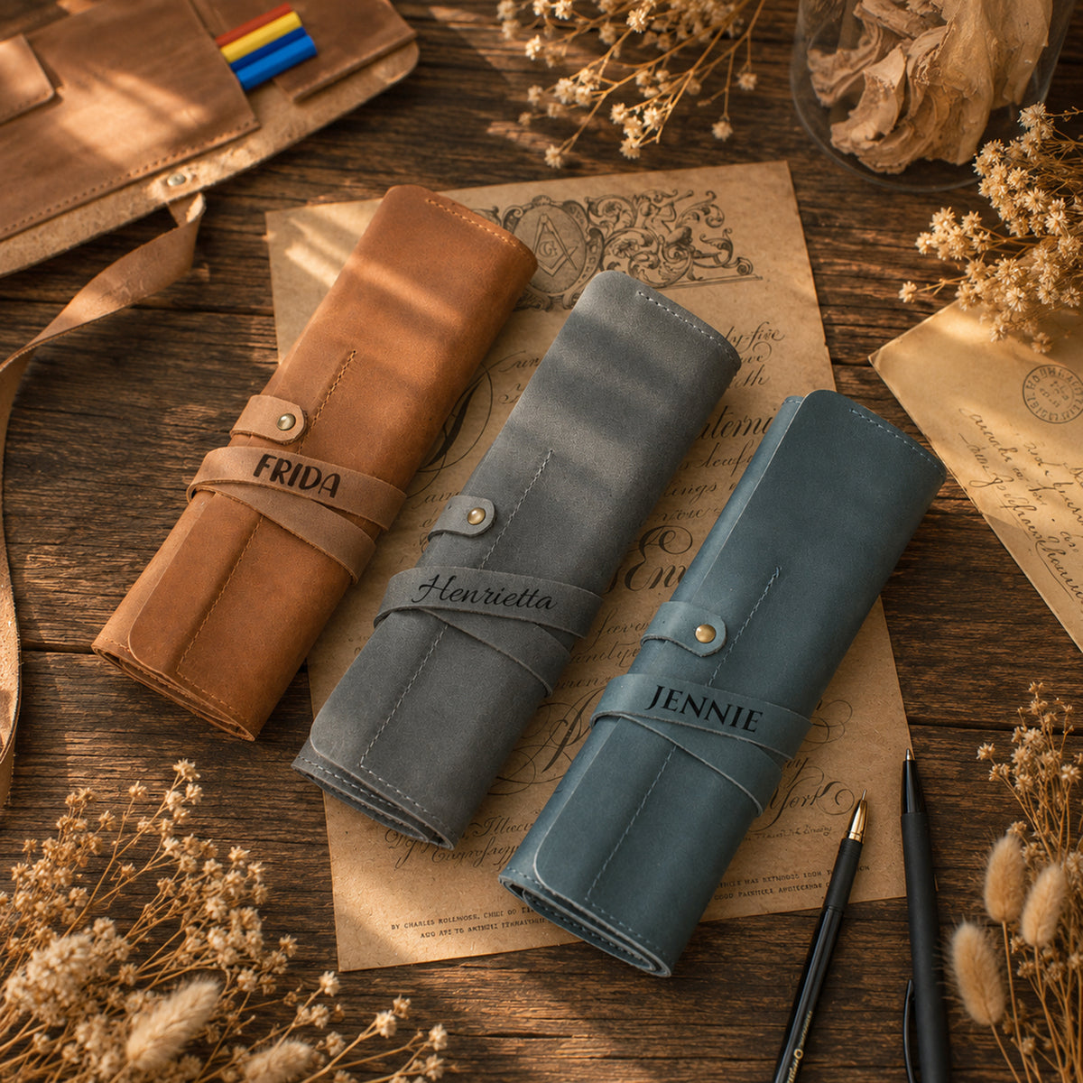 Three rolled leather journal covers in tan, muted blue-gray, and dusty teal are arranged on a rustic wooden surface with vintage paper, dried flowers, and a pen. The covers are personalized with the names FRIDA, Henrietta, and JENNIE on the straps.