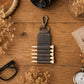 A dark gray leather pencil pouch with a metal clip, shown on a rustic wooden desk surrounded by dried flowers, a camera lens, glasses, and a pocket watch.