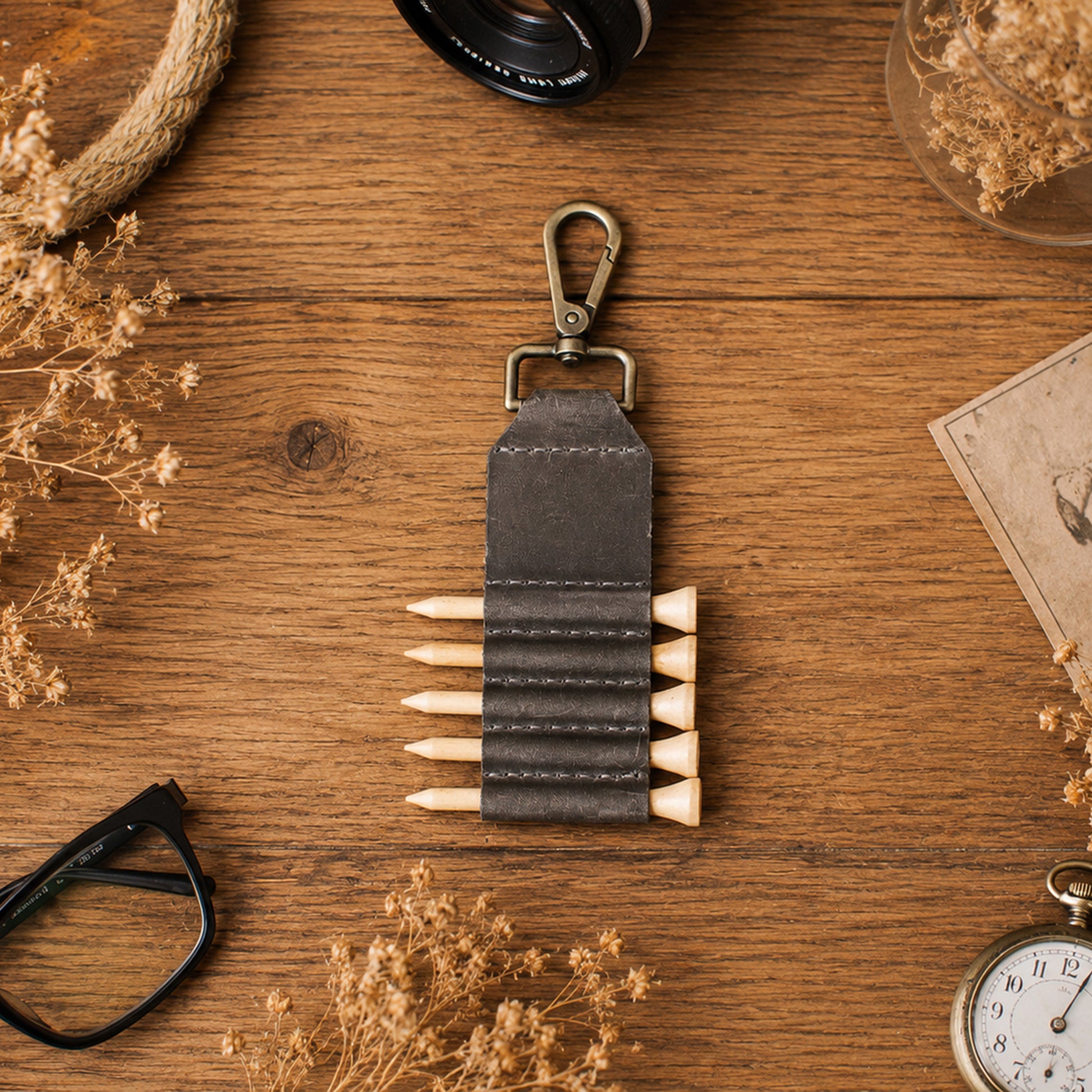 A dark gray leather pencil pouch with a metal clip, shown on a rustic wooden desk surrounded by dried flowers, a camera lens, glasses, and a pocket watch.