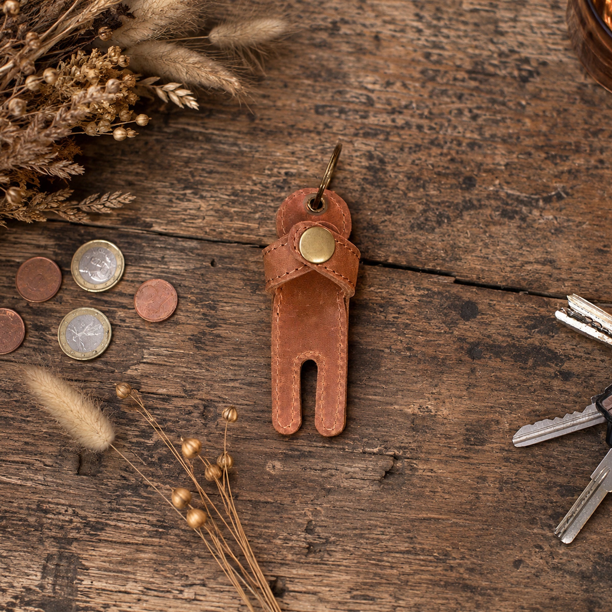 A brown leather golf ball divider keychain with a brass snap button, displayed on a rustic wooden tabletop with keys, coins, and dried grasses.