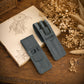 Two personalized blue-gray faux-leather pen holders on a wooden table, styled with an open sketchbook and dried flowers. One holder is open, showing two black pens, and both feature monogrammed pockets with the names 'DH' and 'DANIEL'.