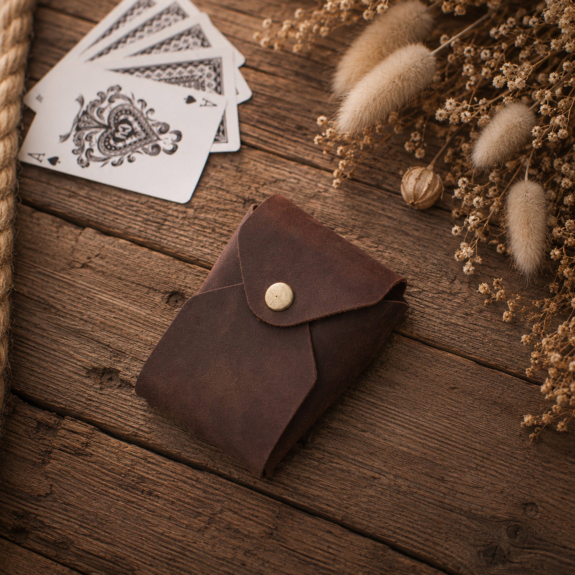 A small brown leather card case with a snap button closure, placed on a rustic wooden surface beside playing cards and dried decorative grasses.