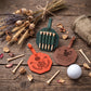 Three personalized leather golf tees and a golf flag set arranged on a rustic wooden table with dried wheat, lavender, and a golf ball. The set includes an orange flag, a green flag with wooden tees, and a brown flag engraved with 'THOMAS', alongside matching leather loops and a tan pouch.