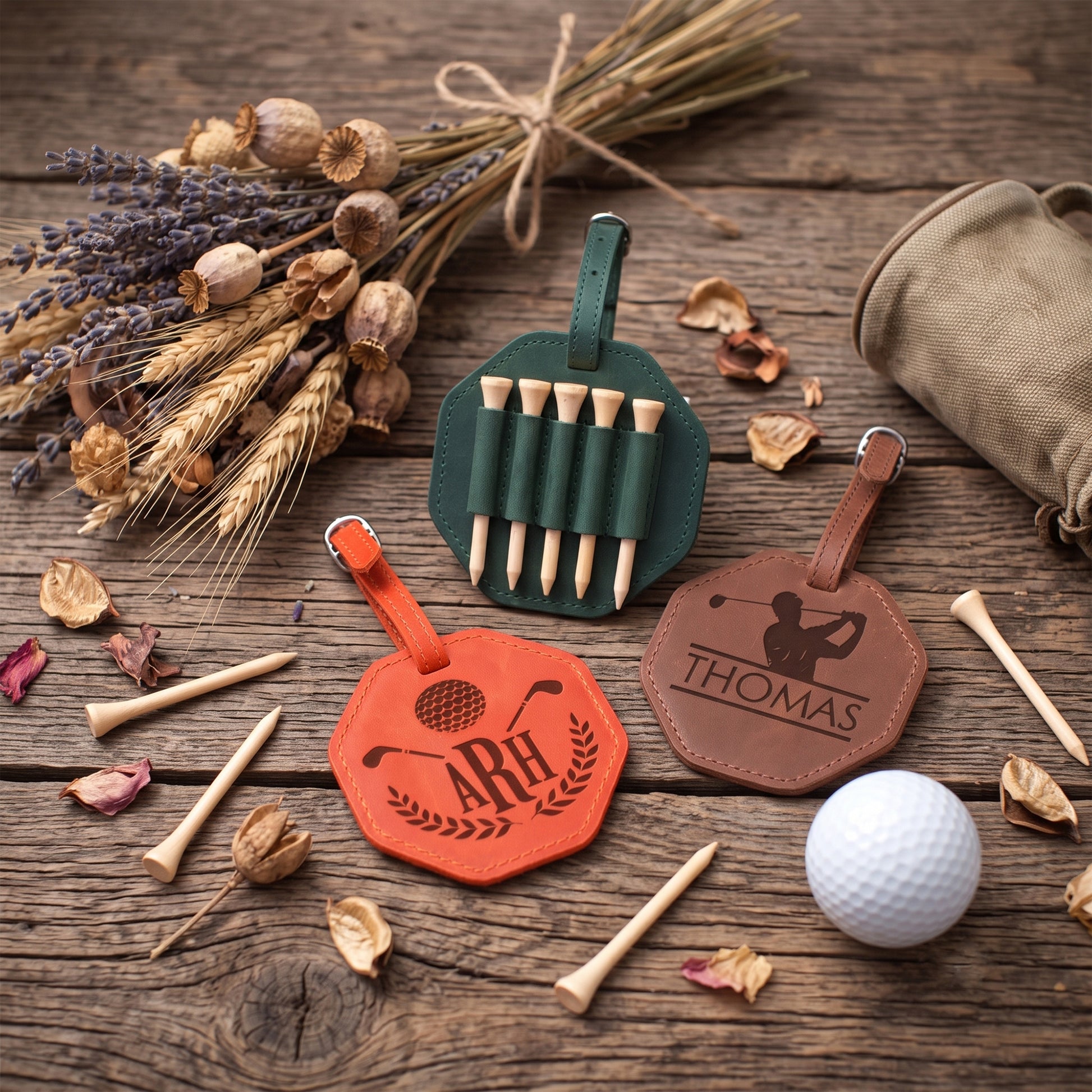 Three personalized leather golf tees and a golf flag set arranged on a rustic wooden table with dried wheat, lavender, and a golf ball. The set includes an orange flag, a green flag with wooden tees, and a brown flag engraved with 'THOMAS', alongside matching leather loops and a tan pouch.