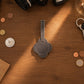 A dark gray leather luggage tag with a floral or clover-like cutout shape and a stitched center, placed on a warm wooden tabletop surrounded by coins, keys, and camera equipment.