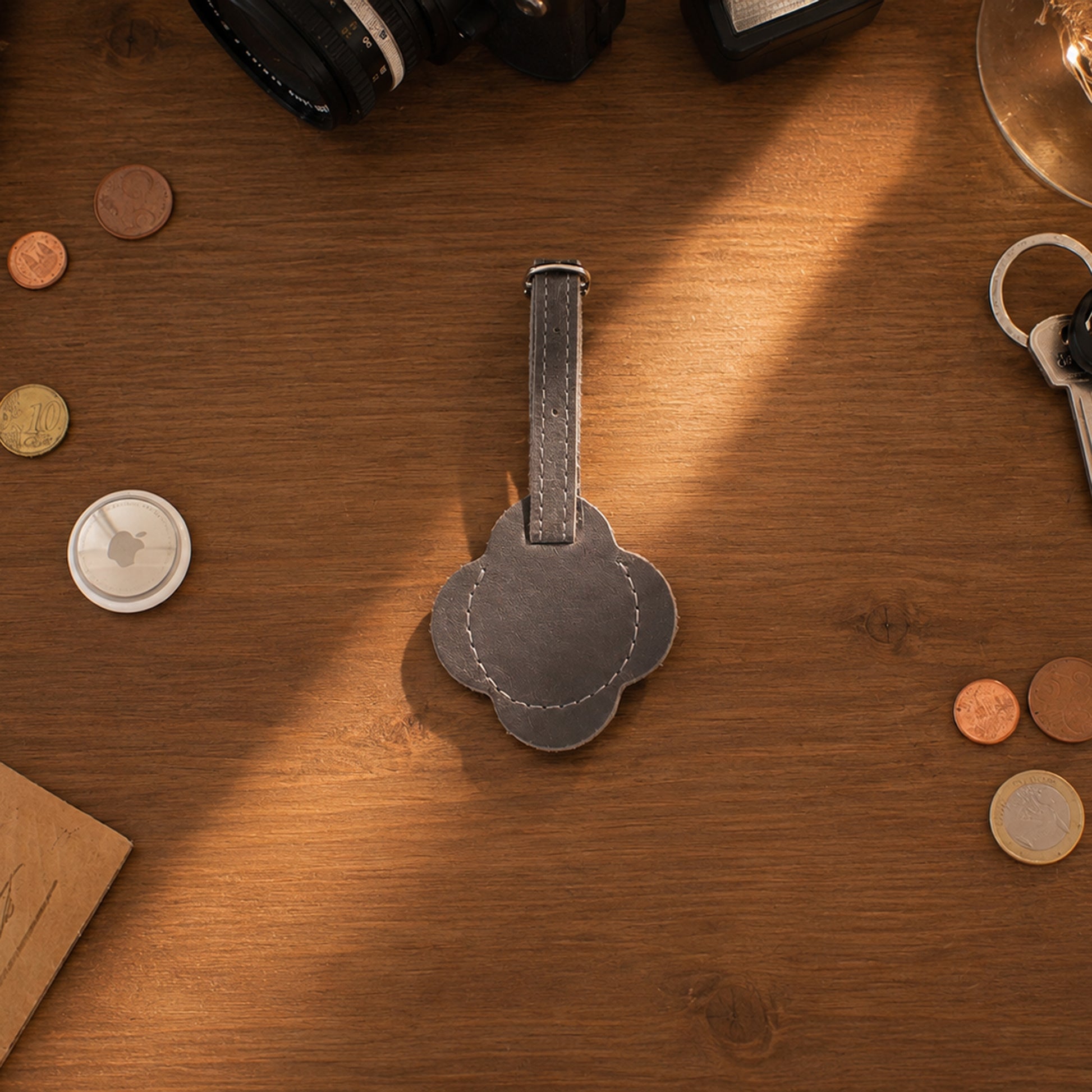 A dark gray leather luggage tag with a floral or clover-like cutout shape and a stitched center, placed on a warm wooden tabletop surrounded by coins, keys, and camera equipment.
