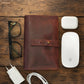 Burgundy leather cable organizer, glasses, white Apple products on a wooden surface