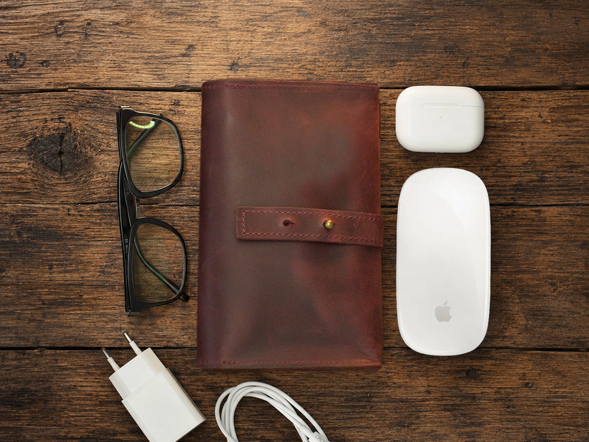 Burgundy leather cable organizer, glasses, white Apple products on a wooden surface