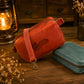 A reddish-brown leather crossbody bag with a black script logo reading 'Charlotte' sits on a rustic wooden table next to a teal pouch. A lit lantern and dried flowers decorate the scene.