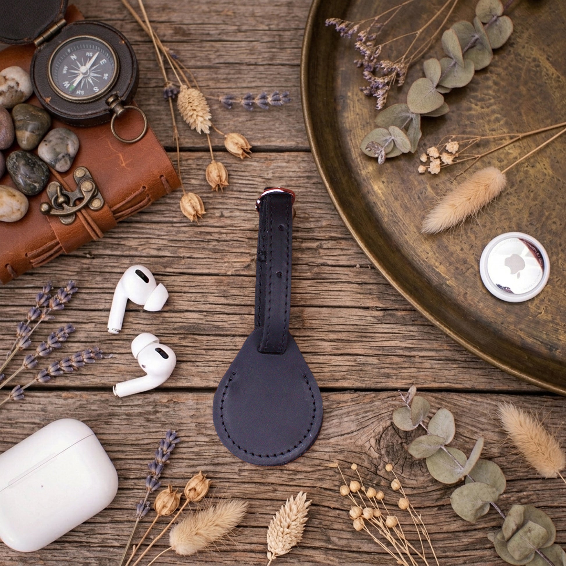 A dark navy leather keychain or bag charm with a stitched oval patch and a metal key ring, displayed on a rustic wooden table surrounded by travel-themed props including a leather wallet, compass, AirPods, and dried flowers.