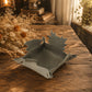 A small gray fabric tray with leaf-shaped flaps, placed on a rustic wooden table in a warm, softly lit room with dried flowers and books in the background.