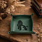 A square green leather dog tray with small side handles, featuring a black silhouette of a dog and the phrase 'Remember who you are' printed inside. The tray is styled on a rustic wooden table with dried flowers and leather accessories in the background.