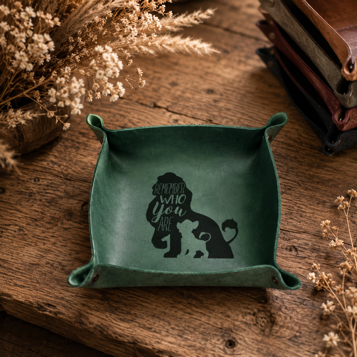 A square green leather dog tray with small side handles, featuring a black silhouette of a dog and the phrase 'Remember who you are' printed inside. The tray is styled on a rustic wooden table with dried flowers and leather accessories in the background.