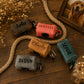 Five personalized dog poop bags in different colors (brown, coral, tan, teal, and gray) with custom names printed on them, arranged on a wooden table with decorative rope, dried flowers, and a picture frame.