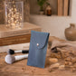 A blue leatherette pouch with a silver snap button, standing on a wooden table surrounded by makeup brushes, dried flowers, and decorative books with warm fairy lights in the background.