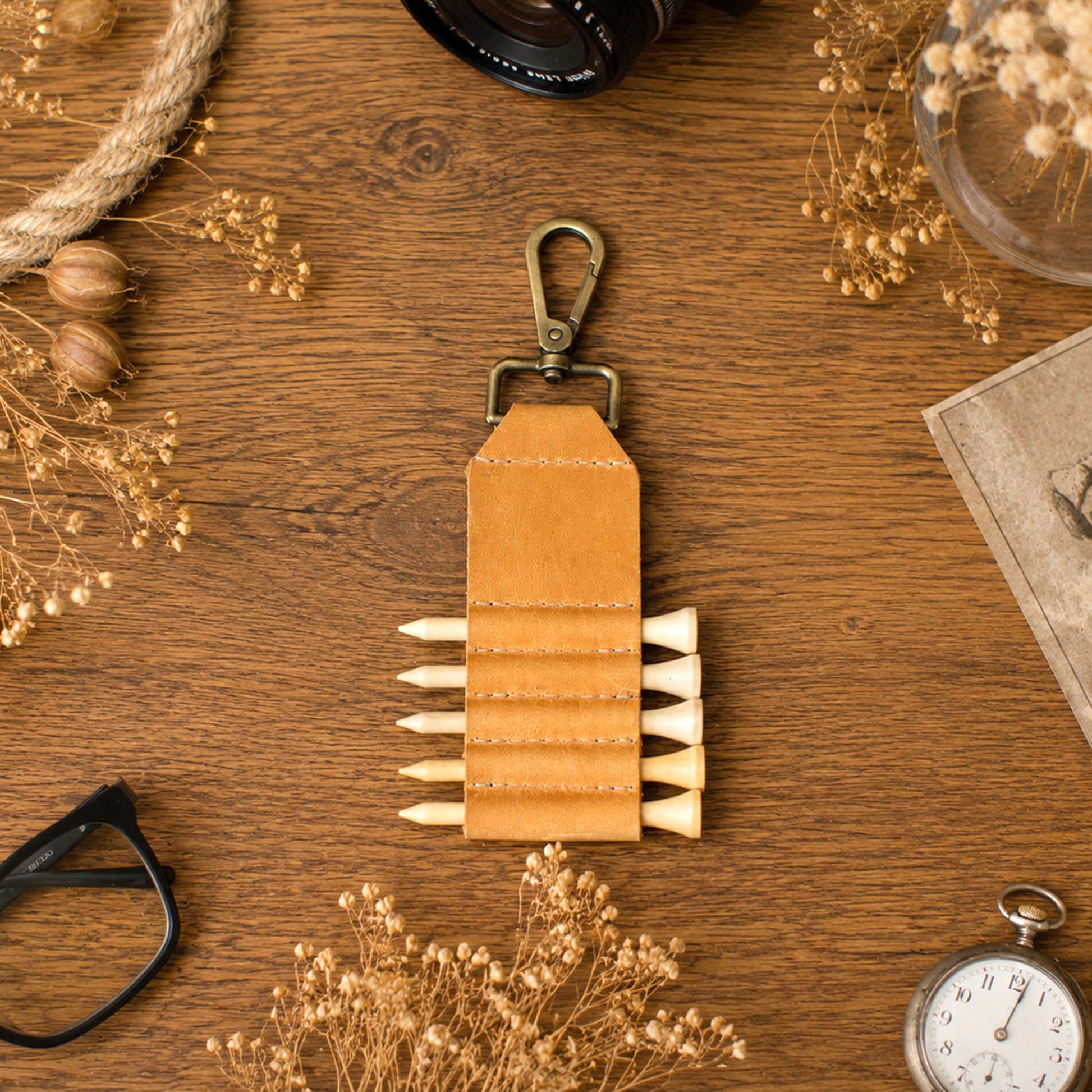 A tan leather key pouch with a brass carabiner and multiple horizontal slots holding white pencils, arranged on a wooden tabletop with dried flowers, glasses, a camera lens, and a pocket watch.