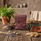 A dark burgundy leather organizer pouch with a cutout window and brass snap buttons, displayed on a rustic wooden table alongside dried flowers, a potted fern, and other leather pouches in green and brown.