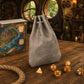 A gray drawstring pouch bag stands on a wooden table, with several translucent amber polyhedral dice scattered in front and a decorative map box to the left. The scene is styled in a fantasy-themed interior with warm lighting and round windows in the background.