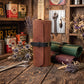 A rolled brown leather tool roll with a black strap and buckle stands on a rustic wooden table, surrounded by vintage-style tools, dried flowers, and other leather rolls in green and burgundy.