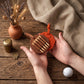 A person's hands holding a brown leather golf club head cover with a bright orange loop, displayed on a rustic wooden table with a golf ball, a small clay pot with dried wheat, and a burlap cloth.