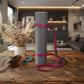 A gray textured yoga mat rolled upright on a wooden table, wrapped with a bright pink strap. Additional rolled straps in brown, tan, blue, and green are arranged around it, with a blurred living room interior in the background.