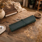 A dark green rolled leather journal or notebook cover rests on a rustic wooden table, surrounded by dried flowers, a vintage book, a small bowl, and white earbuds in a cozy indoor setting.