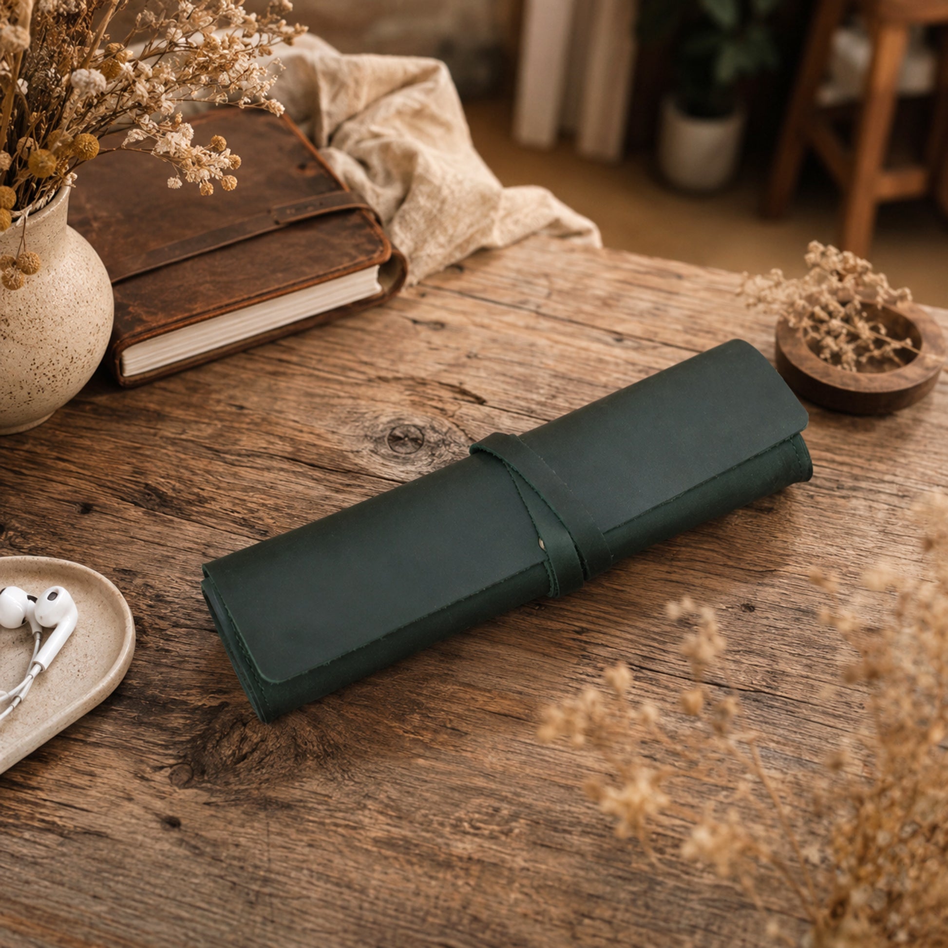 A dark green rolled leather journal or notebook cover rests on a rustic wooden table, surrounded by dried flowers, a vintage book, a small bowl, and white earbuds in a cozy indoor setting.