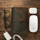 Brown leather cable organizer, glasses, white Apple products on a wooden surface