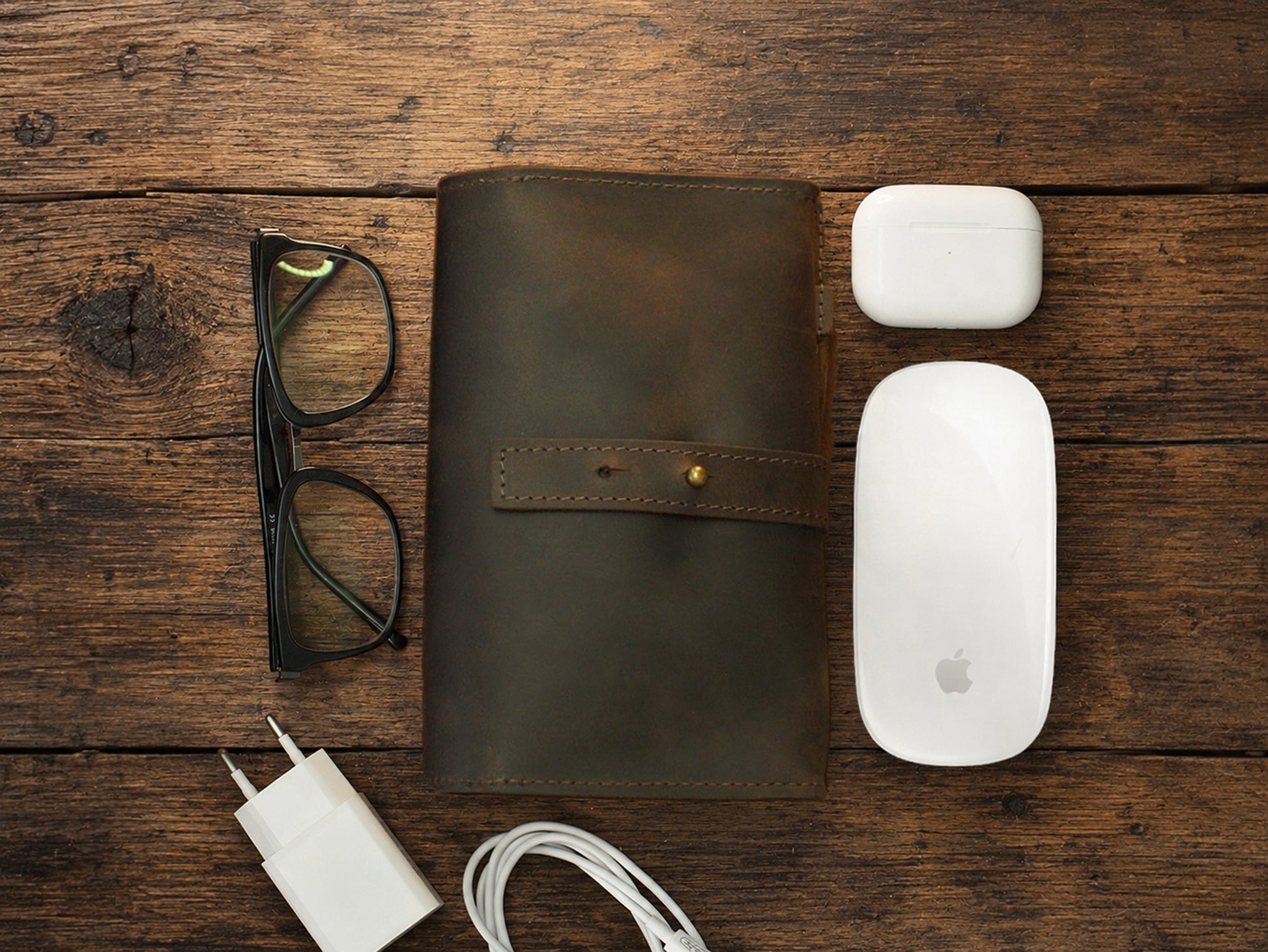 Brown leather cable organizer, glasses, white Apple products on a wooden surface