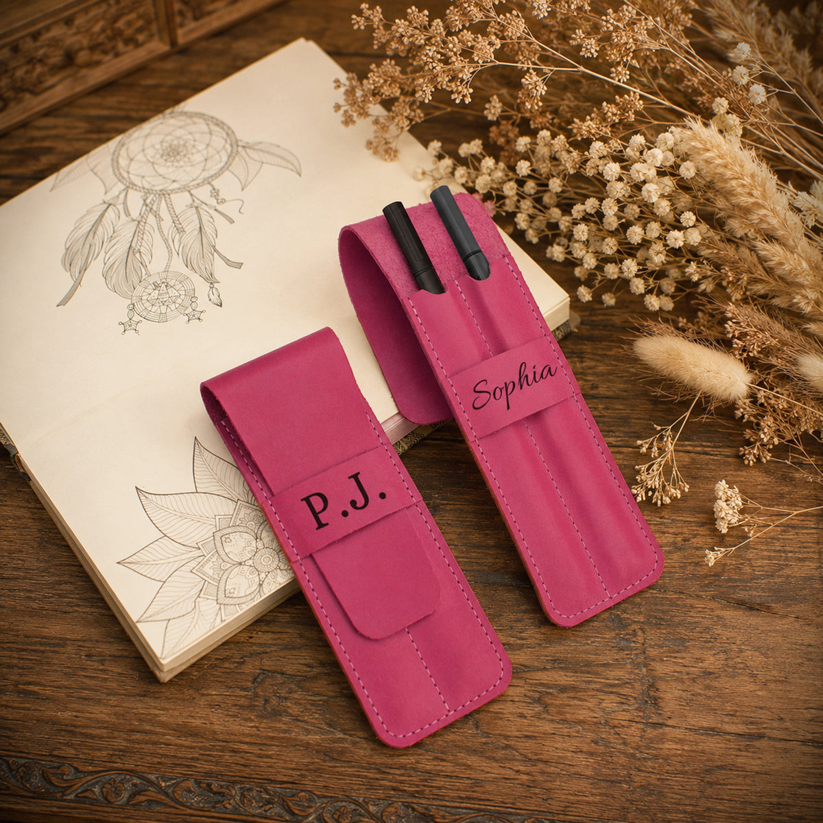 Two pink leather pen cases on a rustic wooden table, styled with an open sketchbook and dried flowers. One case is personalized with 'P.