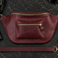 Burgundy leather waist bag with gold zipper on a dark marble surface with accessories.