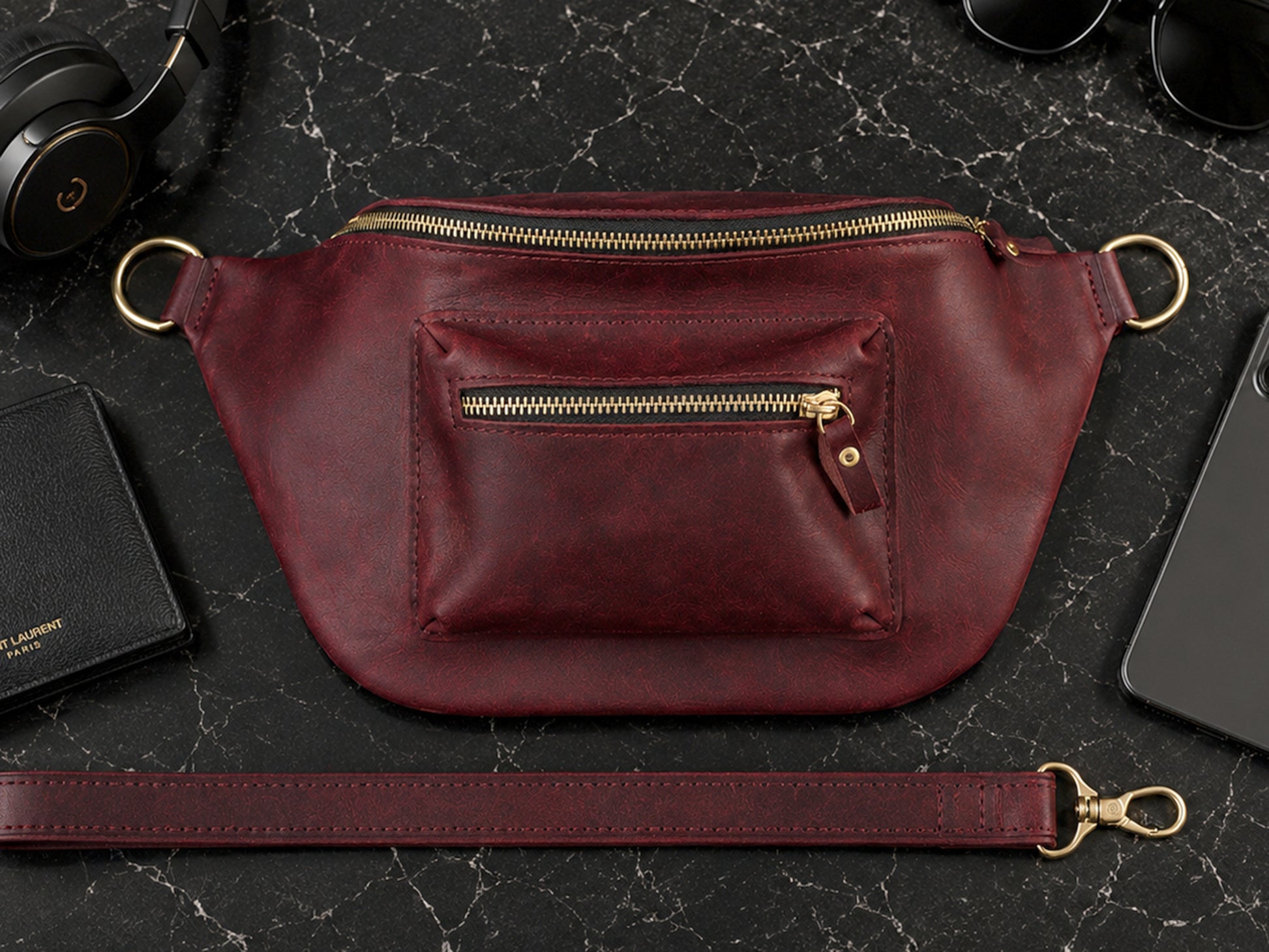 Burgundy leather waist bag with gold zipper on a dark marble surface with accessories.