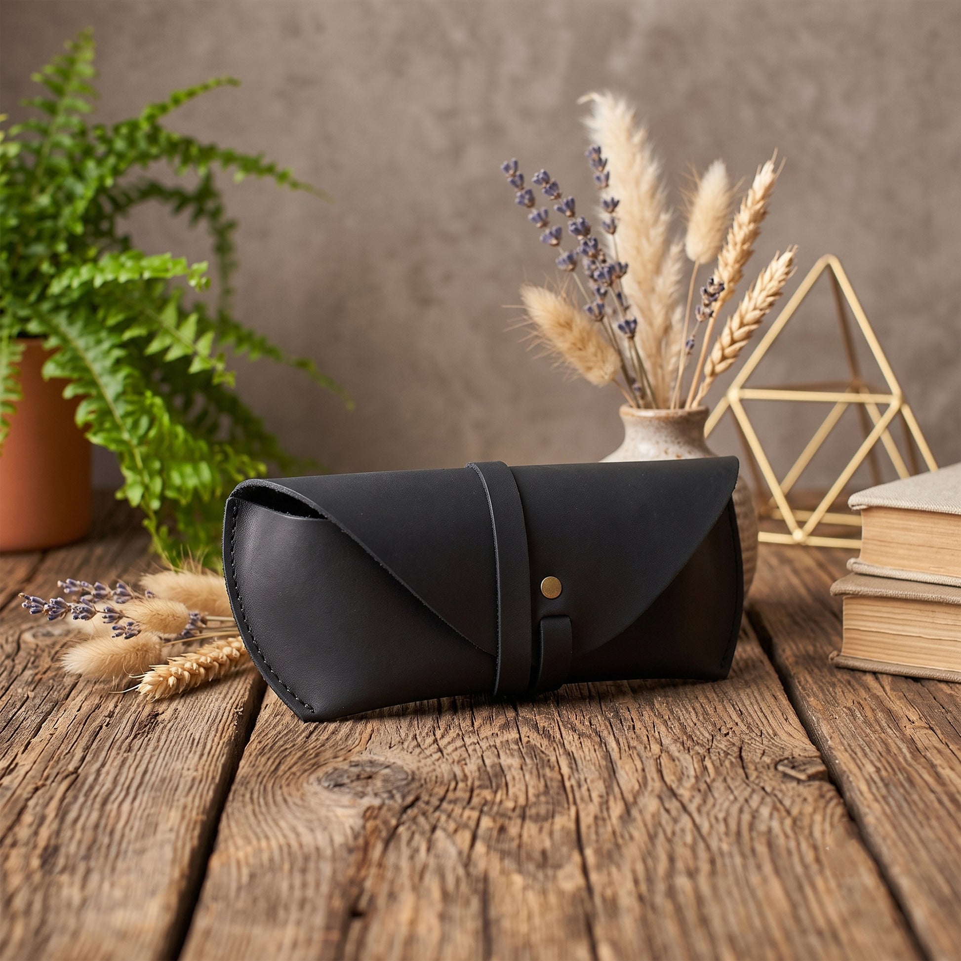 A black leather eyeglasses case with a gold snap button, displayed on a rustic wooden table surrounded by decorative props including a potted fern, dried flowers, books, and a geometric wire frame.