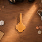 A tan leather luggage tag with a cloud-like outline and a stitched center, displayed on a wooden tabletop surrounded by everyday items like coins, a camera, keys, and a glass.