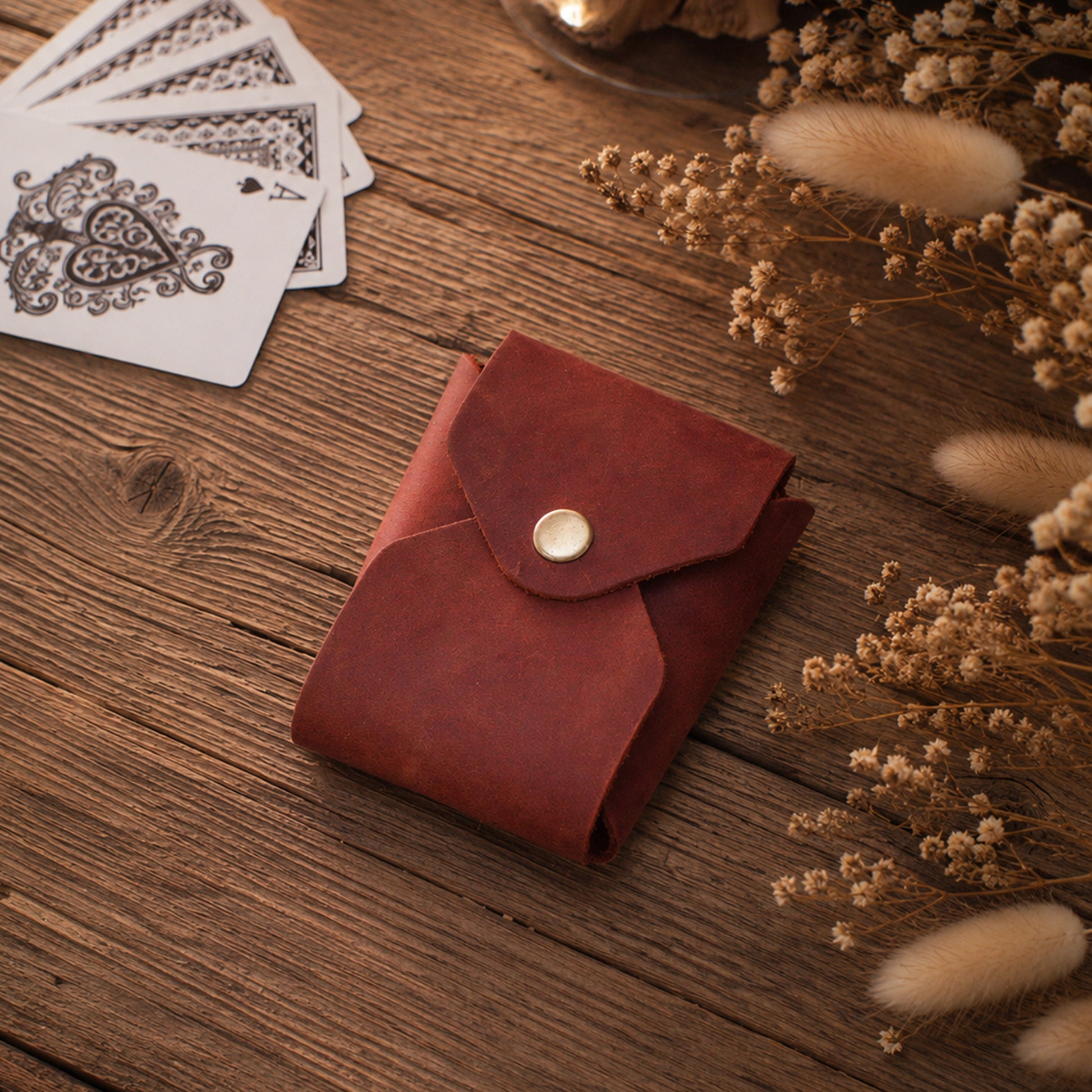 A reddish-brown leather card holder wallet with a snap button closure, styled on a rustic wooden table next to playing cards and dried flowers.