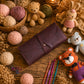 A dark brown leather crocheter's pouch with a strap closure, styled on a rustic wooden table surrounded by yarn balls, crochet hooks, and handmade crocheted toys.