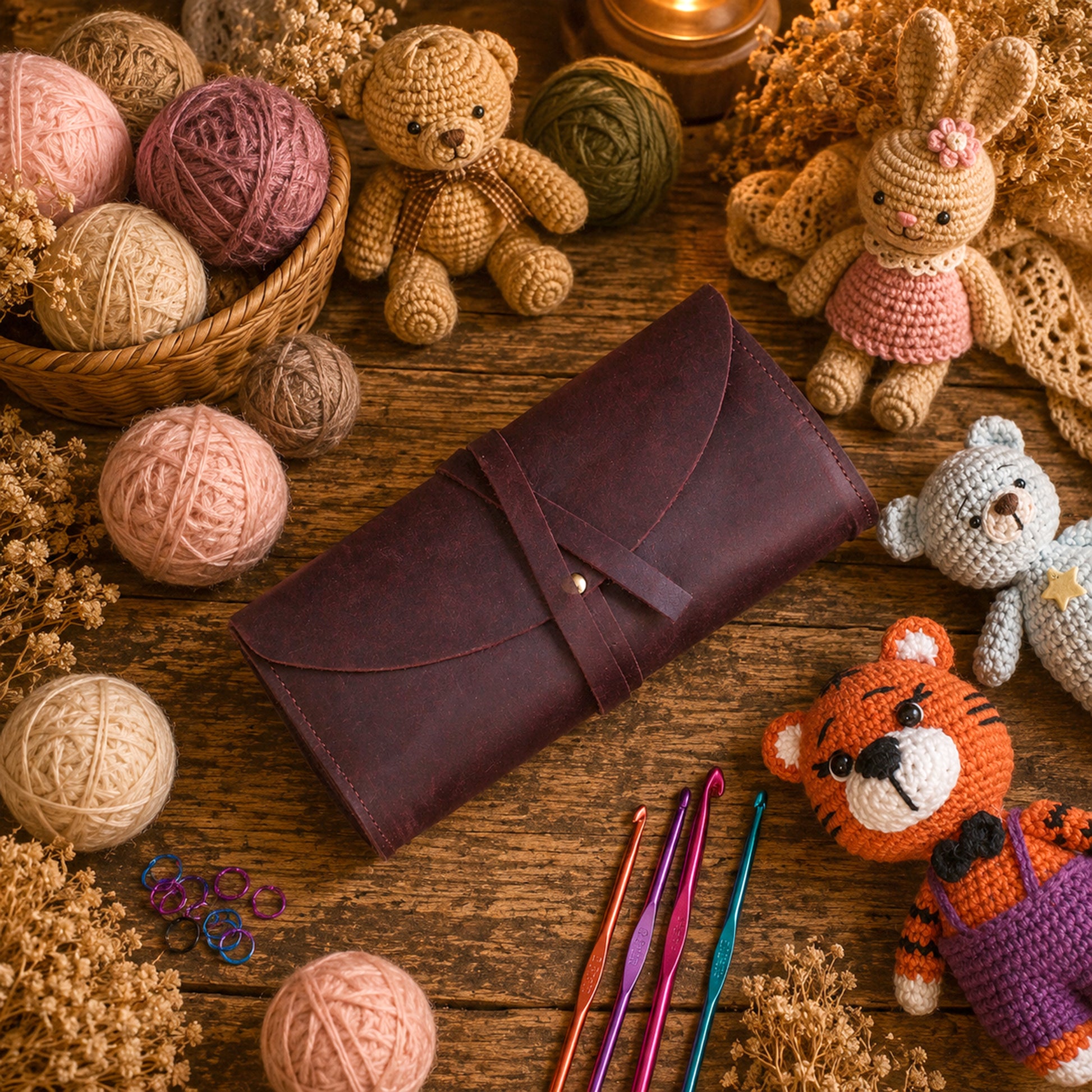 A dark brown leather crocheter's pouch with a strap closure, styled on a rustic wooden table surrounded by yarn balls, crochet hooks, and handmade crocheted toys.