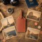 A brown leather luggage tag with a blank nameplate is arranged on a rustic wooden table, surrounded by travel-themed props including vintage postcards of Paris, Rome, Santorini, and Venice, a compass, a camera, coins, and a passport.