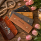 Four personalized leather jewelry pouches in brown, tan, gray, and mustard yellow, arranged on a rustic wooden table with pink tulips and a necklace displayed in the open brown pouch.