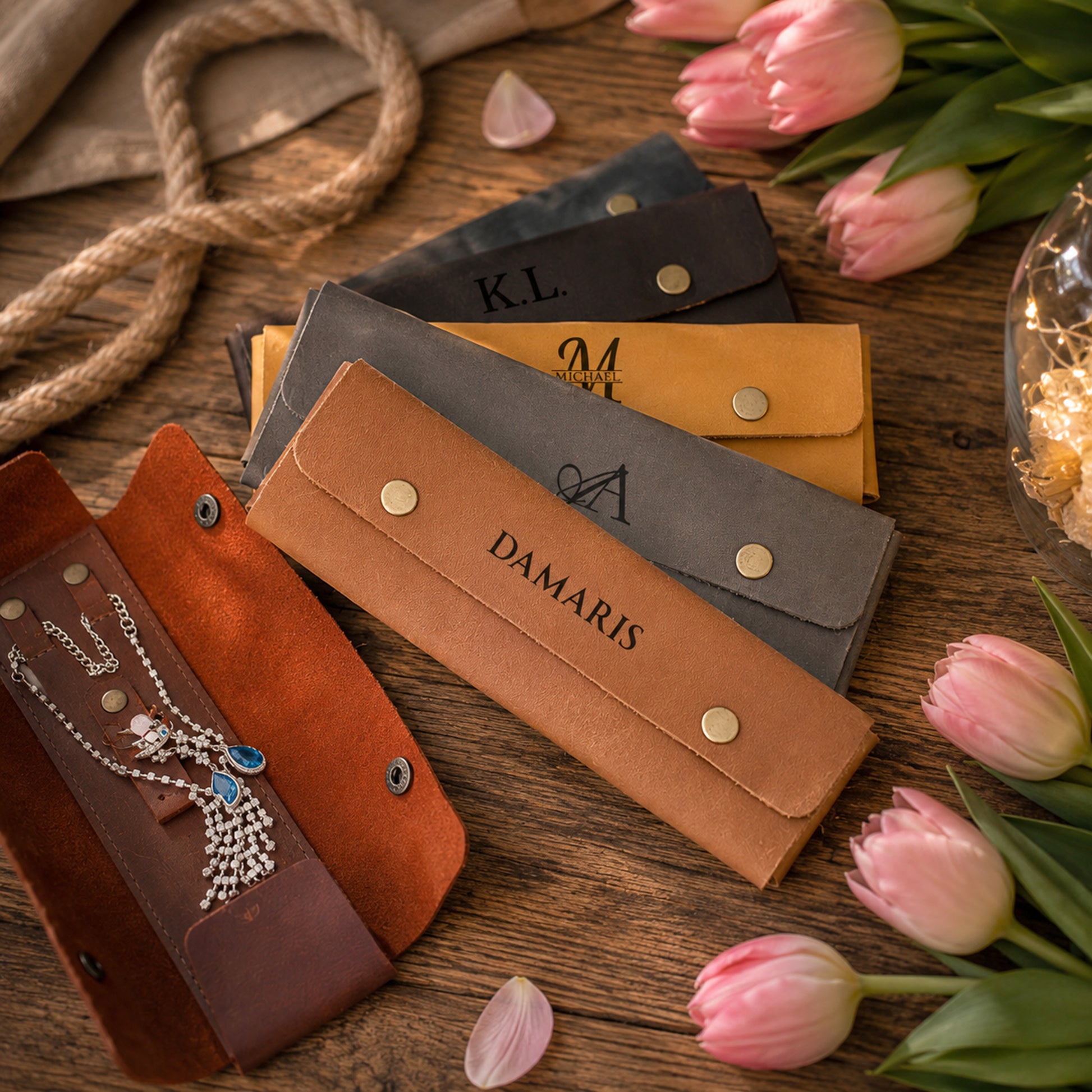 Four personalized leather jewelry pouches in brown, tan, gray, and mustard yellow, arranged on a rustic wooden table with pink tulips and a necklace displayed in the open brown pouch.
