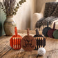Three octagonal golf pencil organizers in orange, brown, and black are arranged on a rustic wooden table, with a white golf ball in front of the center organizer. The background shows a cozy indoor setting with a vase of dried lavender and a blurred armchair.