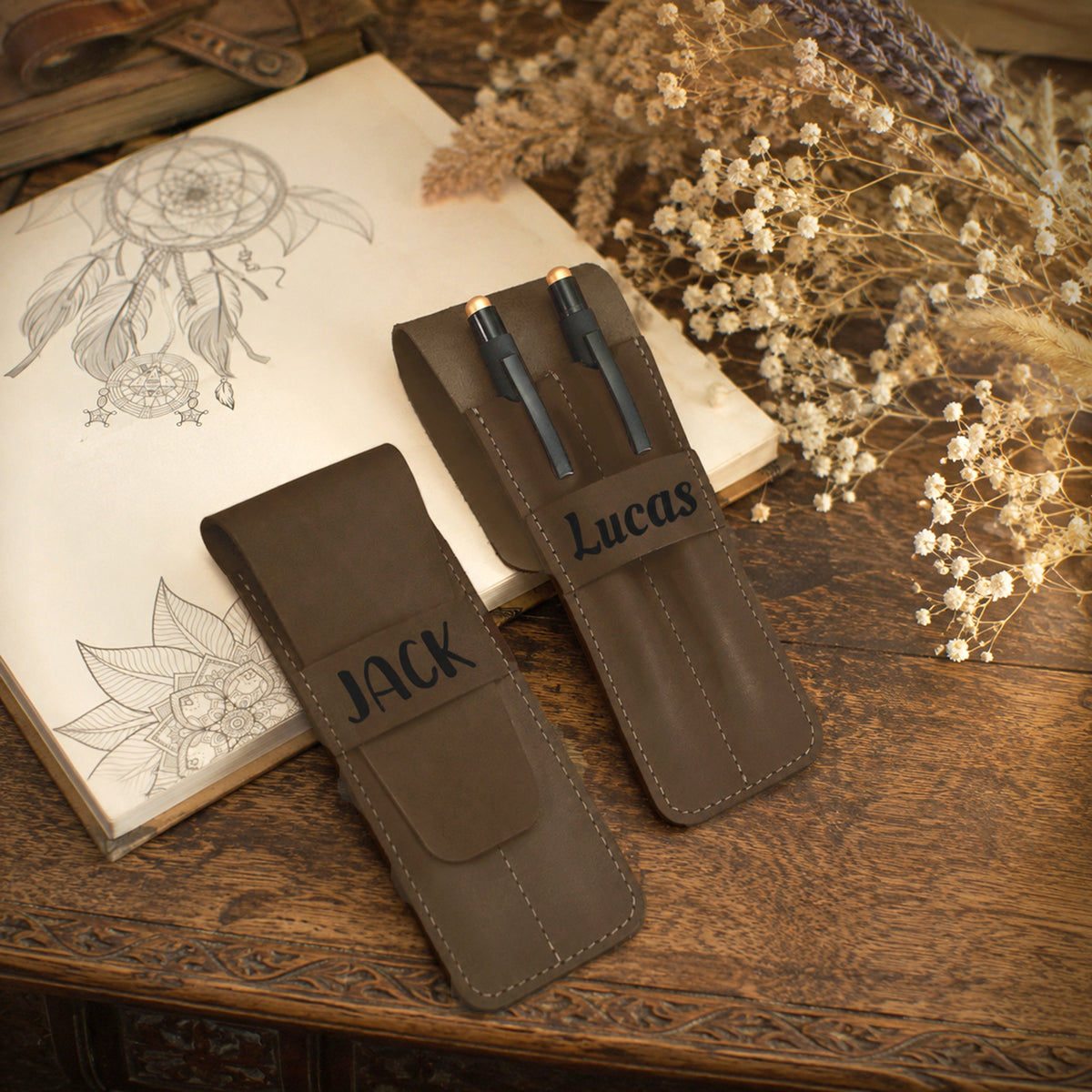 Two dark brown leather pen holders on a wooden table, one labeled 'JACK' and one labeled 'Lucas', styled with an open sketchbook and dried flowers.