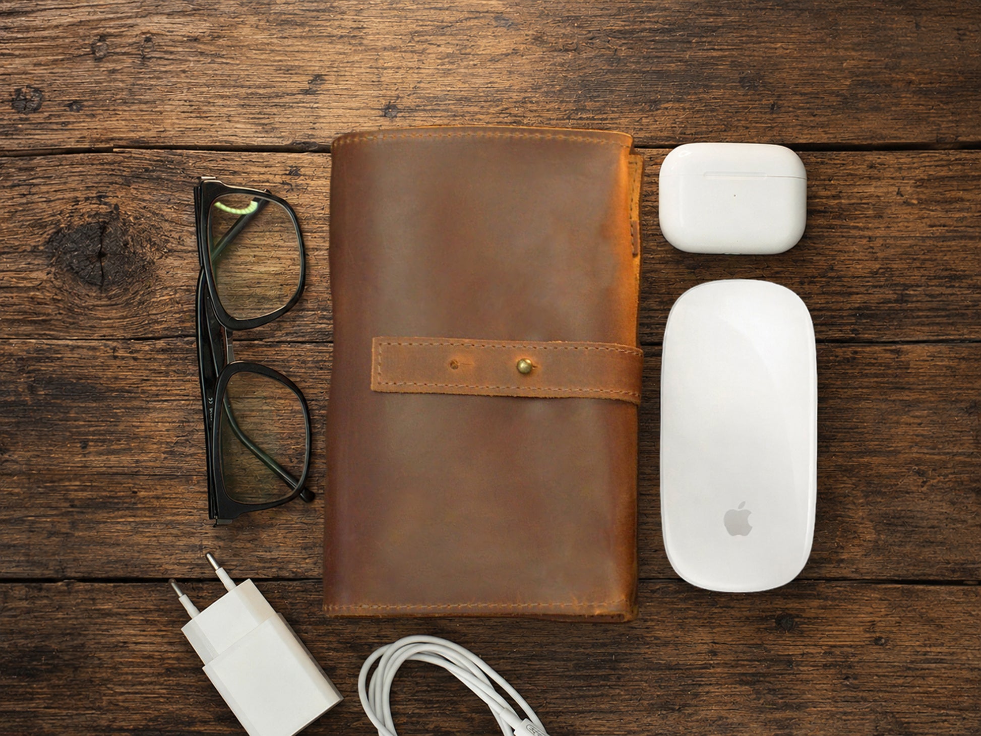 Brown leather cable organizer, glasses, white Apple products on a wooden surface