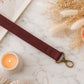 Maroon leather strap with gold clasp on a marble surface with a candle and dried flowers.