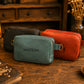 Three small zippered crossbody bags in dark brown, sage green, and rust orange arranged on a rustic wooden table. The front bag is sage green with the name 'MATILDA' printed on the front and a gold-tone ring handle.