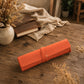 An orange rolled leather journal cover or sleeve with a wrap strap and small gold button, resting on a rustic wooden table beside a book, cloth, and vase of dried flowers.