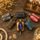 Six small zippered leather pouches in different colors are arranged on a rustic wooden surface with decorative dried flowers, rope, leaves, and a framed photo of a dog. One pouch is personalized with the name 'Tucker' in black script.